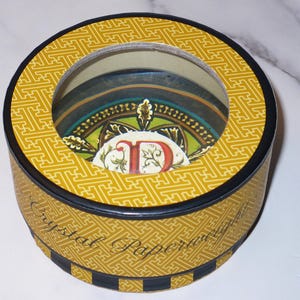 May include: A round decorative box with a clear top, featuring a yellow geometric pattern. The box has a black rim and a decorative interior with a colorful design. The text "Crystal Paper" is visible on the side.