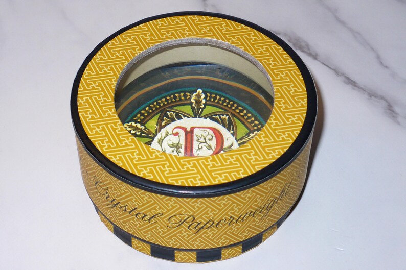 May include: A round decorative box with a clear top, featuring a yellow geometric pattern. The box has a black rim and a decorative interior with a colorful design. The text "Crystal Paper" is visible on the side.