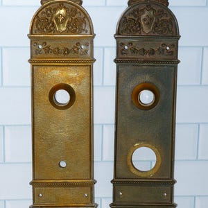May include: Two antique brass door plates with ornate detailing. Each plate features a circular opening and a decorative top with a fleur-de-lis design. The plates have a rectangular shape and are mounted on a white tiled surface.