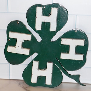 May include: A green metal cloverleaf sign with four white "H" letters, likely for a 4-H club. The sign has a distressed, vintage look, with visible wear and tear. The background is a white tile wall.