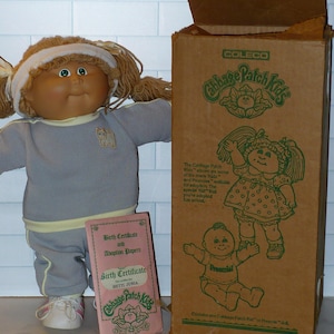 May include: A Cabbage Patch Kids doll wearing a gray outfit with a cat graphic, white shoes, and a headband. A pink birth certificate and adoption papers are next to the doll. A brown box with green graphics and text is on the right.