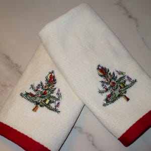 May include: Two white hand towels with a red border. Each towel features an embroidered Christmas tree design with colorful ornaments. The towels are made of a soft, absorbent material, perfect for the holiday season.