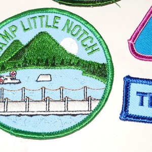 May include: Embroidered patches with various designs. One round patch features a lake scene with mountains and the text "CAMP LITTLE NOTCH". Other patches include a triangle with "cool fun" and a rectangle with "TENDER".