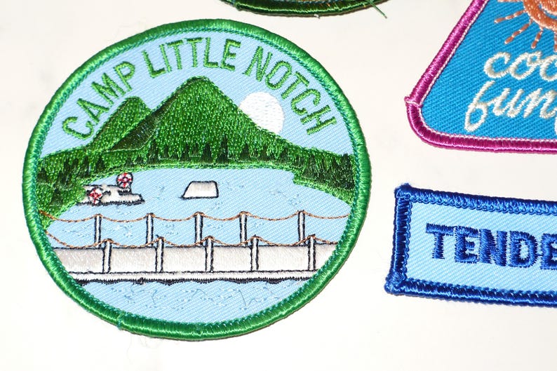 May include: Embroidered patches with various designs. One round patch features a lake scene with mountains and the text "CAMP LITTLE NOTCH". Other patches include a triangle with "cool fun" and a rectangle with "TENDER".