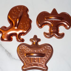 May include: Three copper-colored cookie cutters in the shapes of a lion, a fleur-de-lis, and a crown. The lion cutter has a handle, and the crown cutter has a detailed design. The cutters are on a white marble surface.