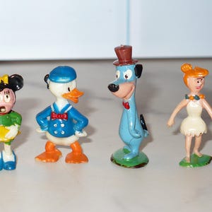 May include: A set of four small plastic character figurines standing in a row. The figures include a mouse in a green outfit, a white duck in a blue sailor suit, a tall blue dog wearing a brown top hat, and a woman with orange hair in a white dress.