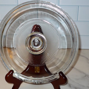 May include: A clear glass lid with a ridged design and a silver knob. The lid is resting on a dark brown wooden stand. The lid is circular and appears to be for a pot or pan. The background is a white tile wall.
