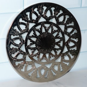 May include: Silver metal trivet with a decorative, intricate, circular design. The design features a central flower with a circular center and a repeating pattern of leaves and swirls.