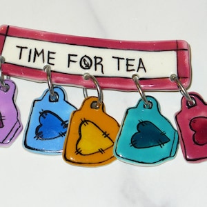 May include: A decorative ceramic piece with the text "TIME FOR TEA". It features a pink-bordered white rectangle with the text, and five heart-shaped tags in purple, blue, yellow, teal, and pink, each with a heart design.