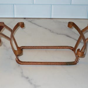 May include: Copper-colored metal wine bottle holder with two wooden accents. The holder has a rectangular shape with a coiled wire base and curved supports. The wooden accents are a dark brown color.