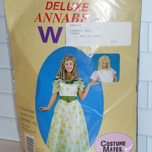 May include: A package for a Deluxe Annabel wig. The package is tan with the word "W" in purple. The package contains a picture of a woman wearing the wig and a dress. The wig is brown. The package is from Costume Mates.