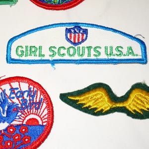 Może przedstawiać: Kolekcja haftowanych naszywek, w tym naszywka Girl Scouts U.S.A. z niebieską obw&oacute;dką i zielonym tekstem. Inne naszywki przedstawiają niebieskiego ptaka, wsch&oacute;d słońca i parę ż&oacute;łtych skrzydeł.