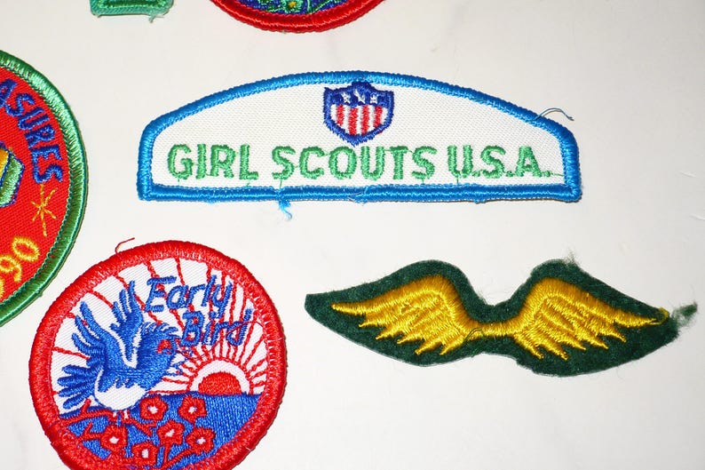 Może przedstawiać: Kolekcja haftowanych naszywek, w tym naszywka Girl Scouts U.S.A. z niebieską obw&oacute;dką i zielonym tekstem. Inne naszywki przedstawiają niebieskiego ptaka, wsch&oacute;d słońca i parę ż&oacute;łtych skrzydeł.