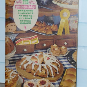 May include: A vintage cookbook titled "The Fleischmann Treasury of Yeast Baking" with various baked goods displayed. The cover features the title in green and pink, with images of bread, rolls, and pastries. A blue and white checkered tablecloth is visible.