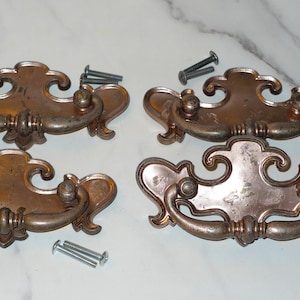 May include: Four antique bronze-toned drawer pulls with ornate, curved designs. Each pull has two small knobs and a central decorative element. Several screws are included, suggesting a hardware set for furniture restoration or a similar project.