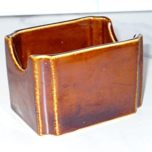 May include: A rectangular brown ceramic napkin holder with a glossy finish. The holder features a U-shaped cutout on the front and back panels, with vertical ribbed detailing along the corners and a smooth, solid-colored glazed surface throughout.