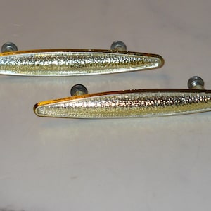 May include: Two gold-toned cabinet pulls with a textured, speckled design. The pulls are oval-shaped with a metallic finish and are mounted with silver screws. These decorative hardware pieces are suitable for furniture restoration or new builds.