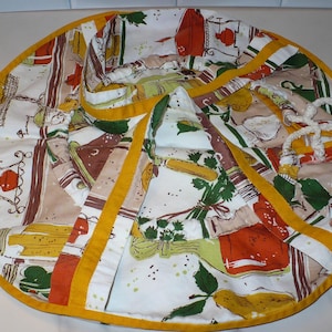 May include: Round, quilted kitchen trivet with a colorful vintage print featuring kitchen utensils, vegetables, and bottles. The trivet has a yellow border and accents, and a white rope loop for hanging. A charming addition to any kitchen.