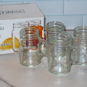 May include: Four clear glass mini drinking jars with handles, displayed with their original box. The box reads "Libbey" and "4 pieces Mini Drinking Jar 4.5 oz/133 ml". The jars are empty and sit on a white surface.