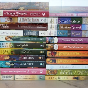 May include: A stack of paperback books with various titles and authors. The spines display titles like "In Sleepy Hollow," "A New Day Rising," and "The Secret." The books are in various colors, including red, blue, and green.