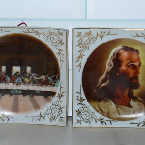 May include: Two square ceramic tiles with religious imagery. One depicts the Last Supper, the other a portrait of Jesus. Both have a white background with gold floral and vine designs around the edges.