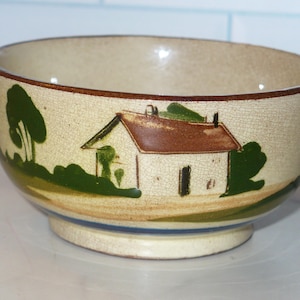 May include: A ceramic bowl with a hand-painted landscape design. The bowl features a cream-colored exterior with a brown rim and a base with blue stripes. The landscape depicts a white house with a brown roof, green trees, and bushes.