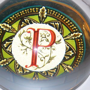 May include: A glass paperweight with a decorative design. The paperweight features a central circle with a red letter "P" and floral accents. The background has green, gold, and dark blue colors, with a clear, rounded glass exterior.