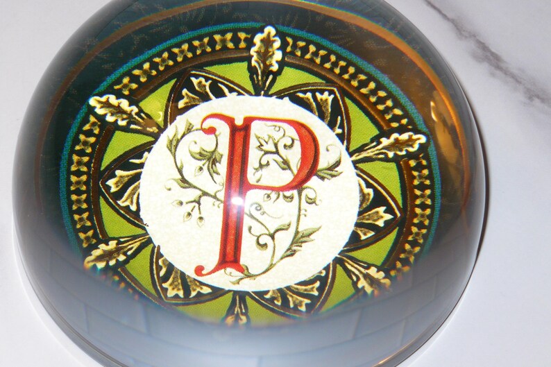May include: A glass paperweight with a decorative design. The paperweight features a central circle with a red letter "P" and floral accents. The background has green, gold, and dark blue colors, with a clear, rounded glass exterior.