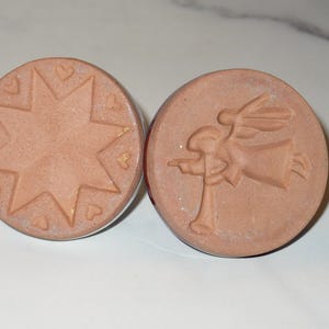 May include: Two round, terracotta-colored stamps. One features a star design with hearts, the other depicts an angel with wings. These stamps are likely used for decorative purposes or crafts.