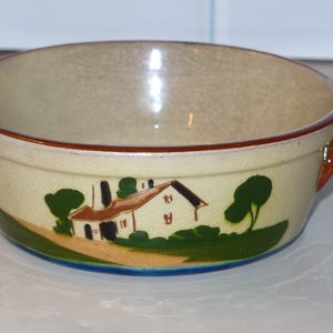 May include: Cream-colored ceramic bowl with two brown handles and a brown rim. The bowl features a painted landscape scene with a house, trees, and a green and blue base. The bowl is likely vintage and suitable for serving.