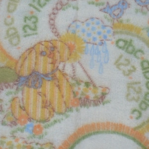 May include: A cream-colored baby blanket with a repeating pattern of a yellow and orange striped bunny, clouds, rainbows, flowers, and the letters "abc" and numbers "123". The blanket has a soft, plush texture.