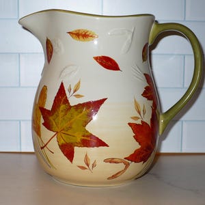 May include: A ceramic pitcher with a cream-colored base and a green handle and rim. The pitcher is decorated with colorful autumn leaves in shades of red, yellow, and orange. The pitcher is designed for serving beverages.