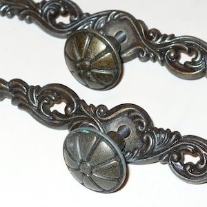 May include: Two antique bronze-toned drawer pulls with ornate, scrolling designs. Each pull features a round, ridged knob and a decorative backplate with intricate details. These vintage hardware pieces are ideal for adding a touch of elegance to furniture.