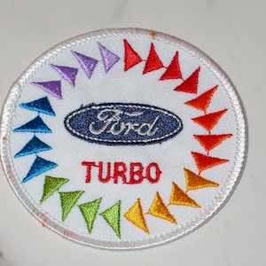 May include: Embroidered patch with a white background, featuring the Ford logo in navy blue and the word "TURBO" in red. A ring of colorful triangles surrounds the logo, with colors ranging from purple to yellow.