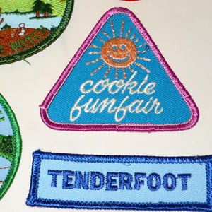 May include: Embroidered patches with various designs. A blue triangular patch features a smiling sun and the words "cookie funfair." A rectangular patch reads "TENDERFOOT" in blue. Other patches are also visible.