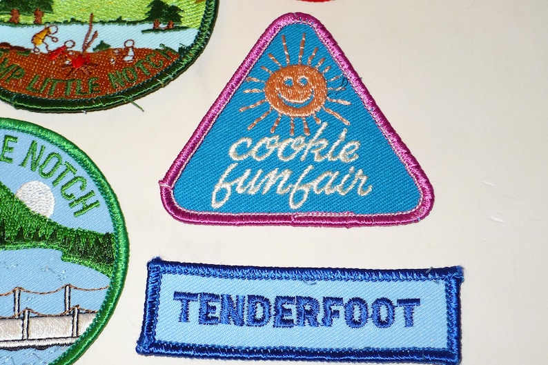 May include: Embroidered patches with various designs. A blue triangular patch features a smiling sun and the words "cookie funfair." A rectangular patch reads "TENDERFOOT" in blue. Other patches are also visible.