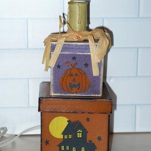 May include: A Halloween-themed decorative light fixture. It features two stacked wooden boxes, one orange with a haunted house design and the other purple with a jack-o'-lantern. A candle-shaped light bulb sits atop the boxes, adding a festive touch.