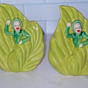 May include: Two green ceramic leaf-shaped planters with a whimsical elf figurine inside each. The elves are wearing green outfits and have red cheeks and smiling faces.