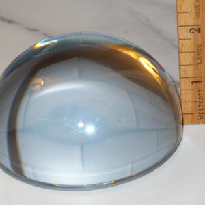 May include: A clear, dome-shaped glass paperweight with a light blue interior. A wooden ruler is positioned next to the paperweight, showing measurements in inches. The paperweight has a reflective surface.