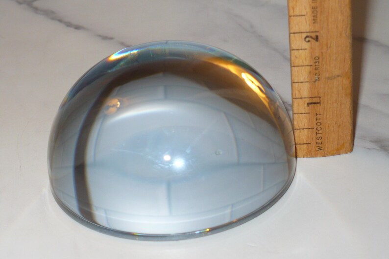 May include: A clear, dome-shaped glass paperweight with a light blue interior. A wooden ruler is positioned next to the paperweight, showing measurements in inches. The paperweight has a reflective surface.