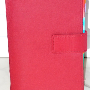 May include: A red fabric organizer with a tab closure. The organizer has a rectangular shape with rounded corners and a stitched border. Colorful divider tabs are visible on the right side.