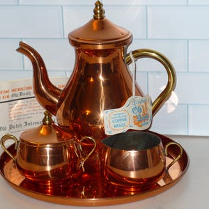 May include: A copper tea set with a teapot, sugar bowl, and creamer on a round tray. The teapot has a gold handle and a tag that reads "Old Dutch Copper Brass". The set is displayed on a white surface with a light blue tiled background.