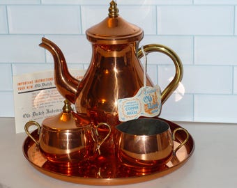May include: A copper tea set with a teapot, sugar bowl, and creamer on a round tray. The teapot has a gold handle and a tag that reads "Old Dutch Copper Brass". The set is displayed on a white surface with a light blue tiled background.
