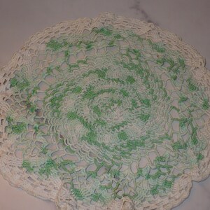 May include: A white crocheted doily with a green and white pattern. The doily is round and has a delicate lace design.