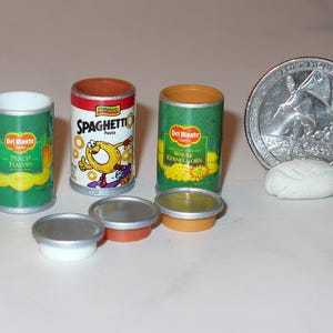 May include: Four miniature cans of canned goods. The cans are green and yellow with white labels. The labels have the text "Del Monte" and the names of the products: "Peach Halves", "Whole Kernel Corn", and "Spaghetti Pasta".