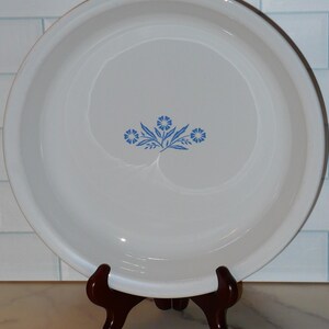 May include: White ceramic pie plate with a blue floral design in the center. The plate has a brown rim and is displayed on a dark brown wooden stand. The background features a white and light blue tile wall.