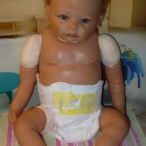 May include: A realistic baby doll with brown hair, blue eyes, and a diaper. The doll is sitting on a pink and green striped blanket.