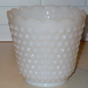 May include: A white hobnail glass vase with a scalloped rim. The vase has a cylindrical shape and a textured surface covered in small, raised bumps. The vase is likely used for decorative purposes or to hold flowers.