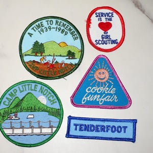 May include: A collection of embroidered patches with various designs and text. One patch reads "A Time to Remember 1939-1989." Another says "Service is the of Girl Scouting." Other patches include "Camp Little Notch," "cookie funfair," and "Tenderfoot."