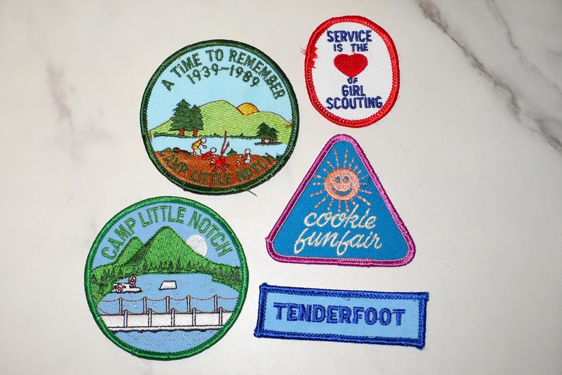 May include: A collection of embroidered patches with various designs and text. One patch reads "A Time to Remember 1939-1989." Another says "Service is the of Girl Scouting." Other patches include "Camp Little Notch," "cookie funfair," and "Tenderfoot."